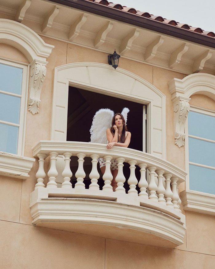 Millie Bobby Brown wearing angel wings, leaning on a balcony railing of a beige building, looking contemplative.