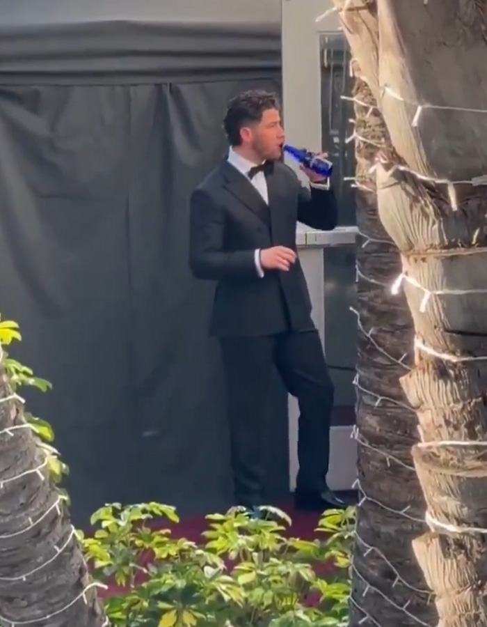 Nick Jonas in a tuxedo standing outdoors, drinking from a blue bottle during the Golden Globes 2026 event.