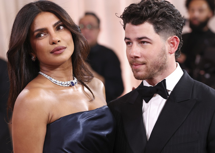 Nick Jonas in a black tuxedo at an event, accompanied by a woman in a navy blue gown and diamond necklace.