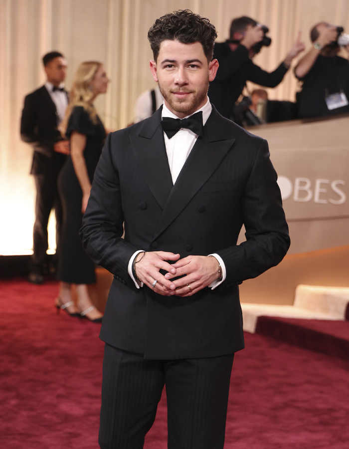 Nick Jonas dressed in black tuxedo walking at Golden Globes 2026 with photographers capturing moments on the red carpet.