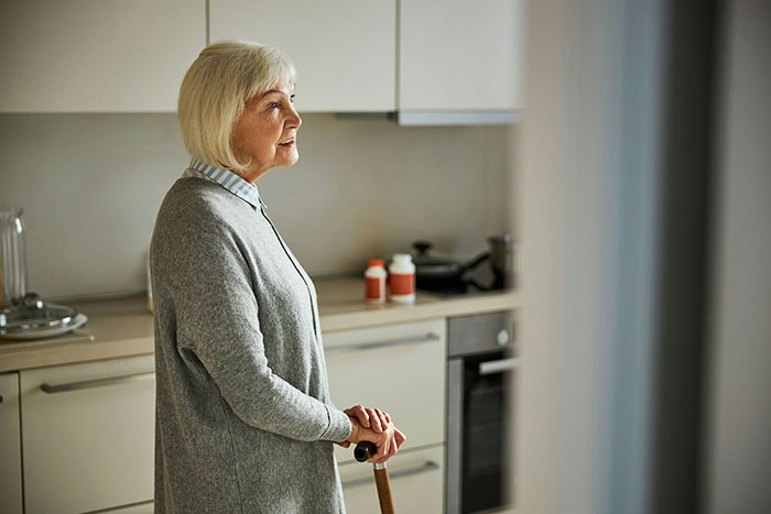 Older woman standing in kitchen with a cane, reflecting on family and retirement plan expectations.