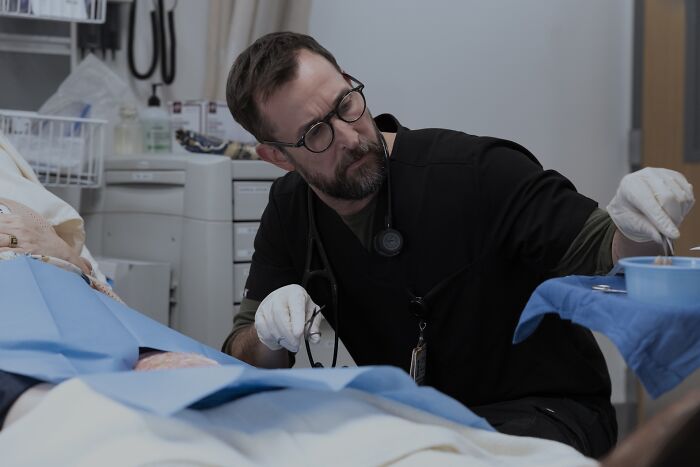 Doctor in black scrubs and glasses performing a medical procedure, referencing The Pitt Season 2 tragic true story.