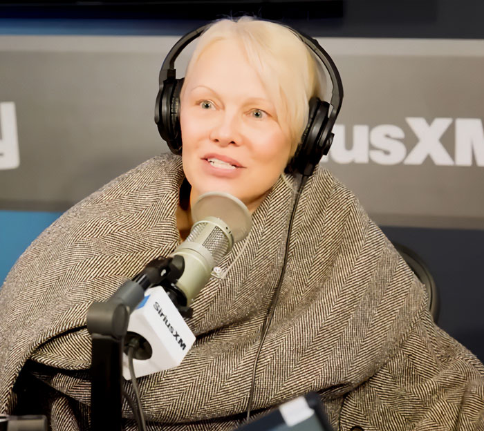 Pamela Anderson speaking into a studio microphone wearing headphones, discussing Seth Rogen Golden Globes apology.