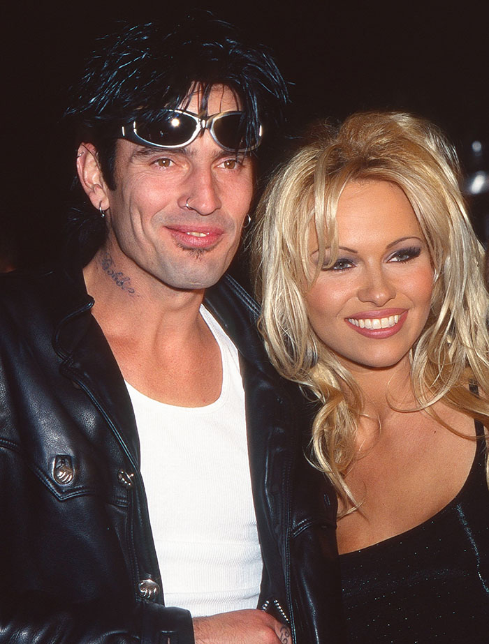 Pamela Anderson smiling in a black dress with a man wearing sunglasses and a leather jacket at an event.