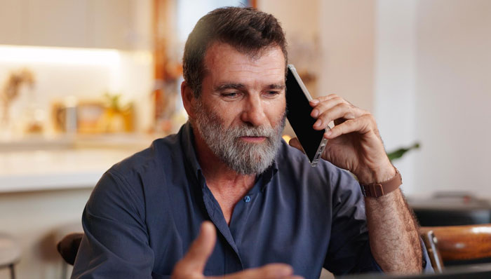 Middle-aged man with beard talking on phone, appearing serious while discussing parents disowning adult worker daughter. Middle-aged man with beard talking on phone, appearing serious while discussing parents disowning adult worker daughter.