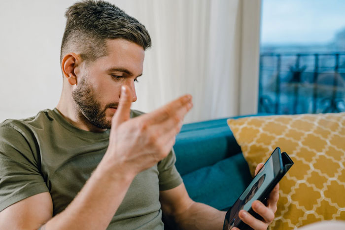 Man sitting on couch looking frustrated while holding a tablet, depicting parents trying to guilt-trip couple into having kids.