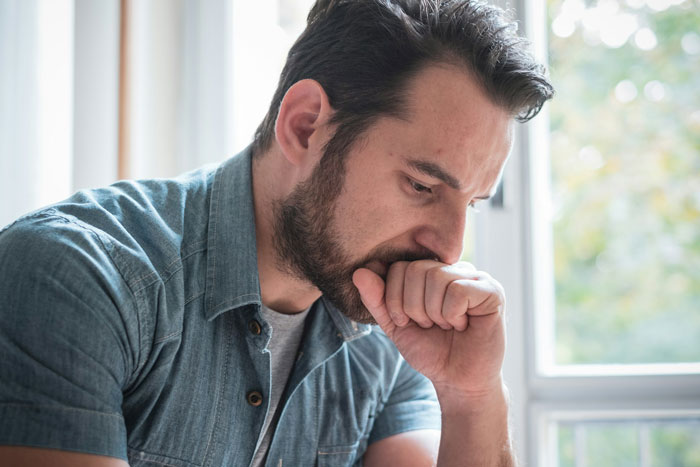 Man sitting by window, looking worried and contemplative while dealing with parents trying to guilt-trip couple about having kids.