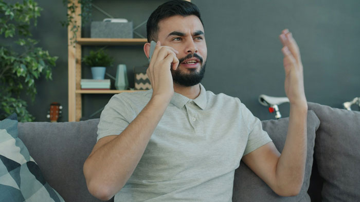 Man talking on phone looking frustrated at home, illustrating man&rsquo;s parents trying to guilt-trip couple into having kids.