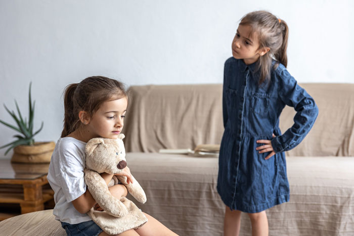 Two young girls in a living room, one looks upset holding a teddy bear, the other stands with a stern expression.