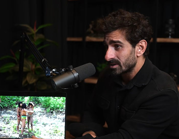 Man speaking into a microphone in a dark room with footage of an uncontacted Amazonian tribe on a laptop screen. Man speaking into a microphone in a dark room with footage of an uncontacted Amazonian tribe on a laptop screen.