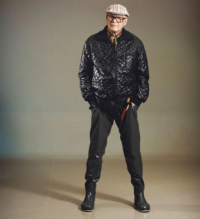 Elderly man wearing a quilted jacket, flat cap, and rain boots posing indoors, evoking strong country memories.