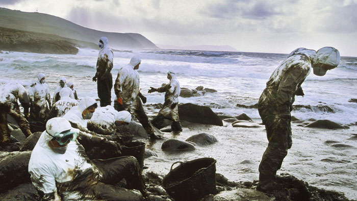 Volunteers in protective suits cleaning an oil spill on a coastline, showing environmental impact in their country.