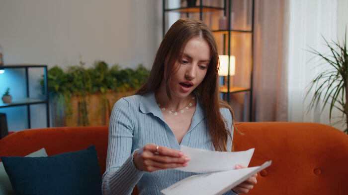 Young woman sitting on orange couch looking shocked while reading papers, illustrating people share wildest ways found out cheating.