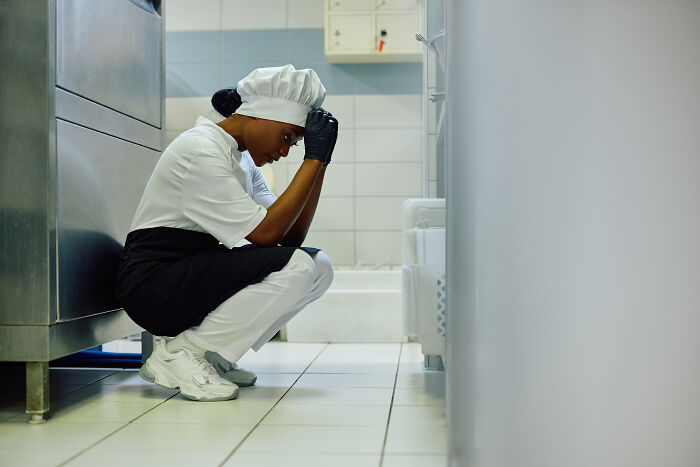 Stressed chef in uniform crouching on kitchen floor, feeling overwhelmed by the worst person they’ve ever met experience.