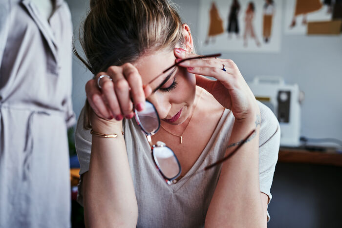 Stressed woman holding glasses in a creative workspace, reflecting on stories about the worst person they've met.