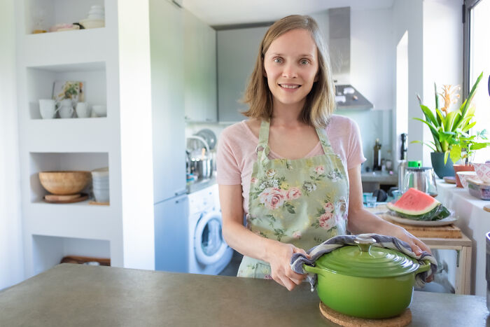 Woman in floral apron holding green pot in bright kitchen, illustrating stories about the worst person they've ever met