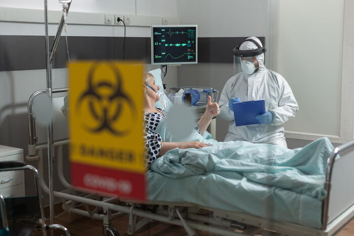 Patient giving thumbs up to healthcare worker in full PPE at bedside with biohazard sign, Moments People Faced