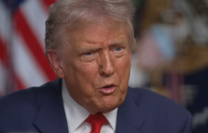 Close-up of older man in navy suit and red tie speaking in interview with American flag background, Moments People Faced