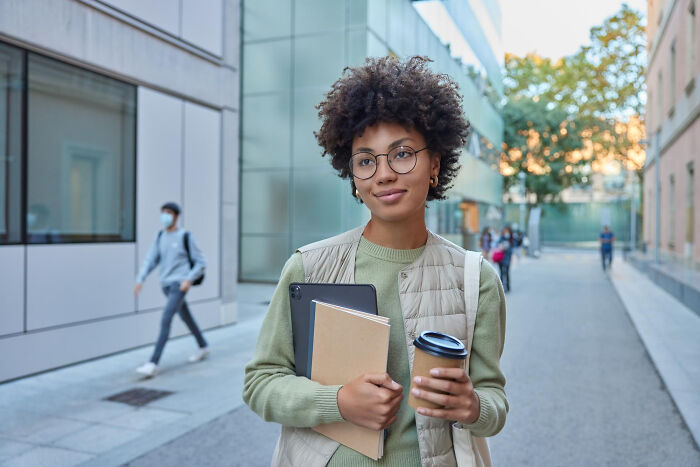 Young woman with glasses holding a tablet, notebook, and coffee cup outdoors, portraying transformation and personal change.