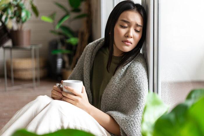 Woman wrapped in a gray shawl holding a cup, looking contemplative near a window, illustrating strange tales of transformation.