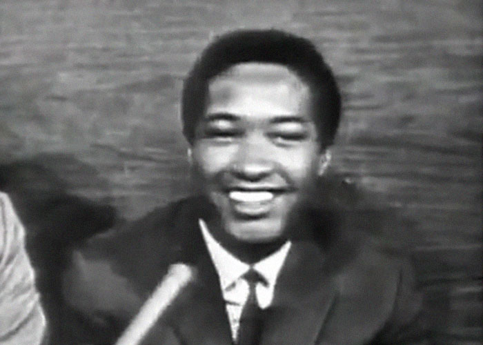 Black and white image of a smiling man in a suit during an interview about celebrity passings still questioned today.
