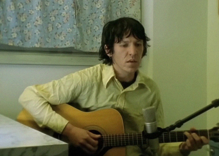 Young man playing acoustic guitar while singing into a microphone, related to celebrity passings topic.