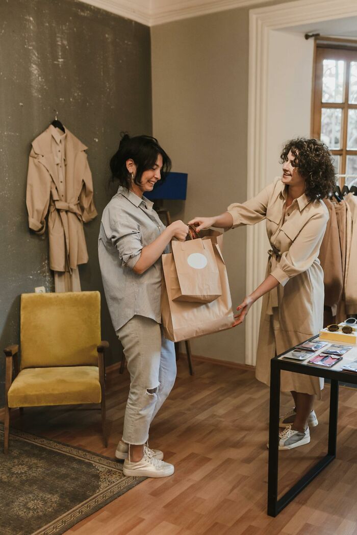 Two women smiling and exchanging shopping bags in a cozy retail space illustrating rich people far from reality.