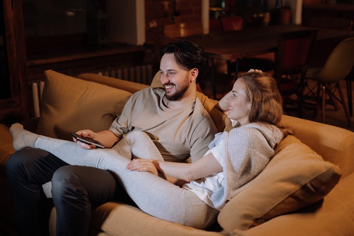 Couple relaxing on couch at home, smiling and watching TV, facing pressure from man&rsquo;s parents about having kids.