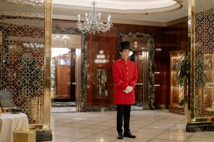 A hotel doorman in a red coat and top hat stands inside a luxurious lobby, illustrating how far from reality rich people can be.