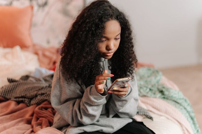 Young woman with curly hair sitting on a bed, anxiously reading accidental text messages on her smartphone.