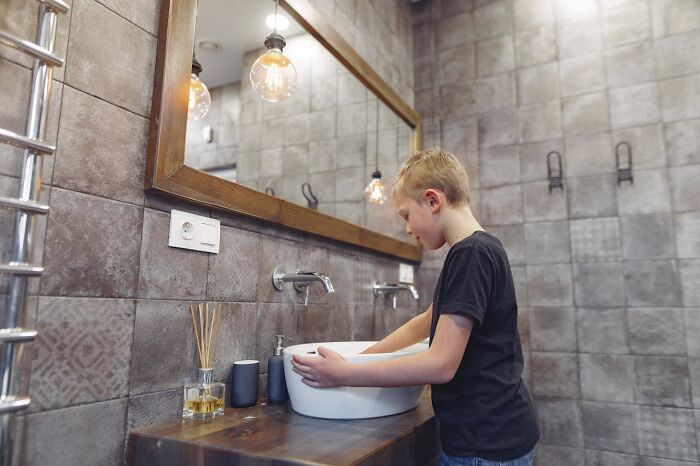 Boy in a modern bathroom washing hands at a sleek sink, highlighting rich people’s disconnect from everyday reality.