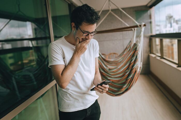 Man in glasses reacting with panic while reading accidental texts on phone near balcony hammock