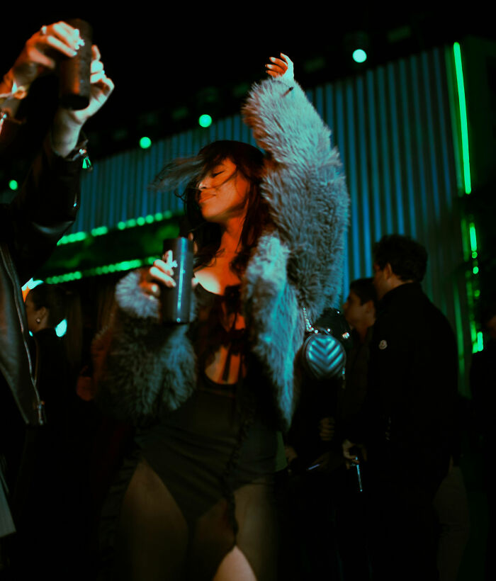Woman dancing with a drink in hand at a nightclub, surrounded by colorful lights and partygoers in a lively atmosphere.