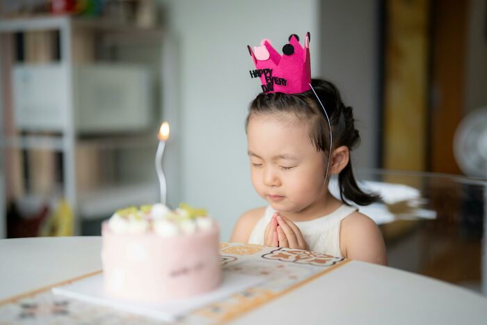 Young girl wearing a birthday crown, closing eyes and praying before blowing out a candle, hinting at family secrets and celebrations.
