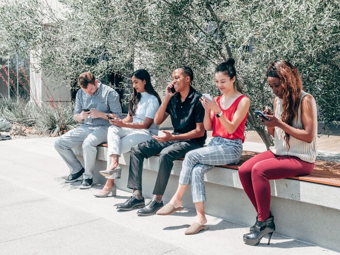 A diverse group of people sitting outdoors on a bench, focused on their phones, illustrating shock after prison experiences.
