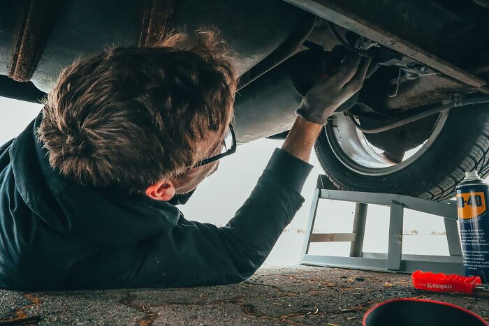 Man in black jacket and gloves inspecting car tire underneath, illustrating rich people out of pocket sayings.