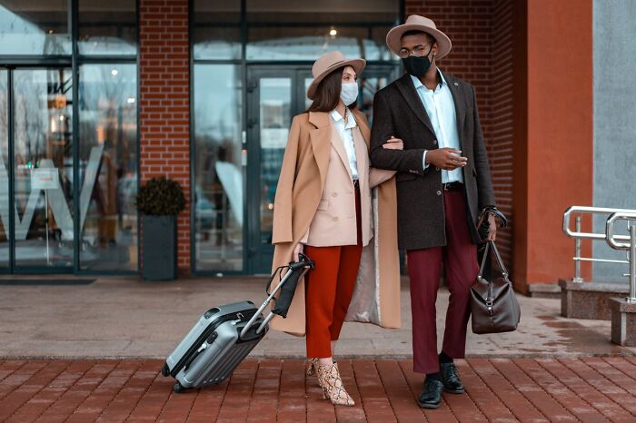 Stylish rich people wearing masks walk with luggage, showcasing how far from reality their luxury lifestyle feels.