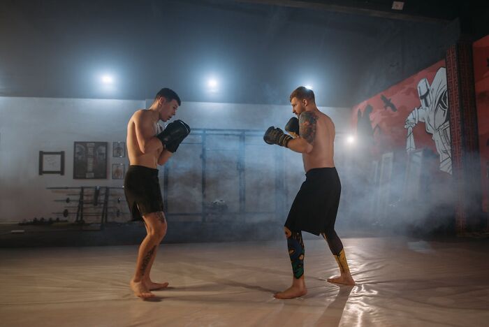 Two shirtless men wearing boxing gloves sparring in a dimly lit gym during a chaotic night shift training session.