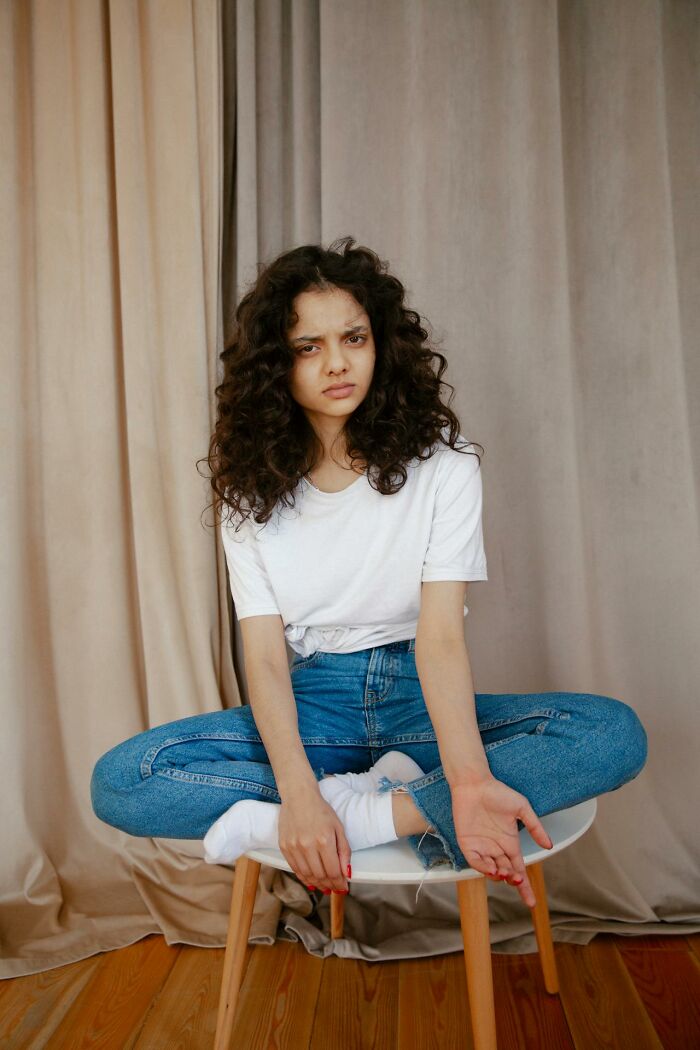 Young woman with curly hair sitting cross-legged on a stool, showing a frustrated expression with rich people disconnect theme.