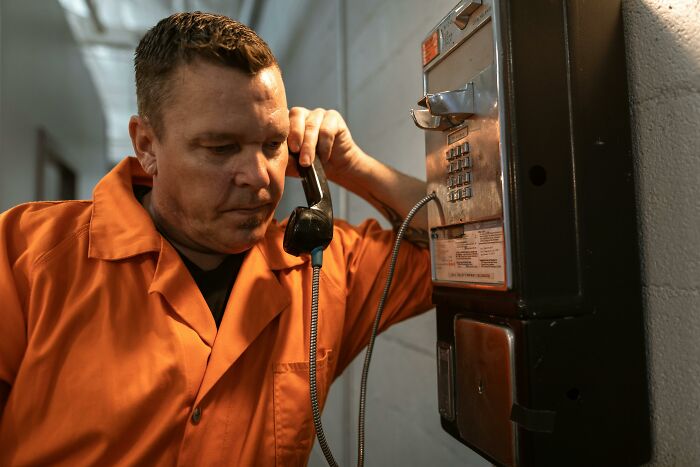 Man in orange prison uniform using a wall phone inside a prison corridor illustrating life in prison misconceptions.
