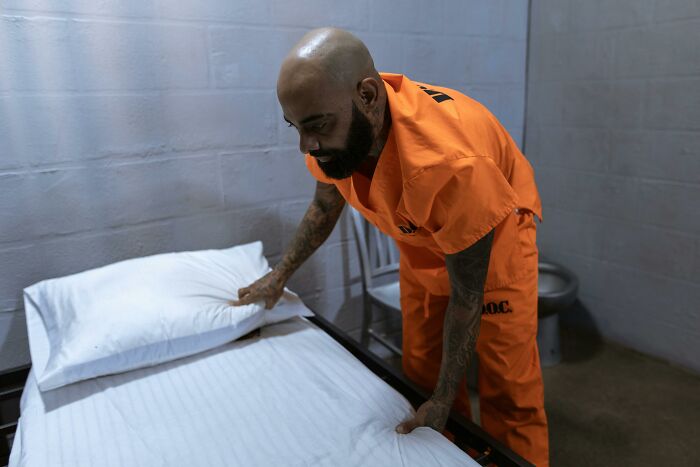 Man in orange prison uniform making a bed inside a cell illustrating life in prison misconceptions.