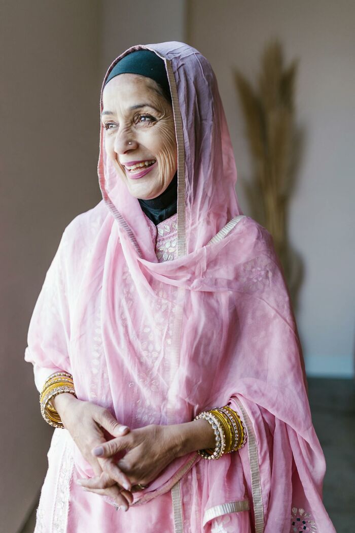 Elderly woman in pink hijab and gold bangles smiling by a window, rained frogs anecdote illustration