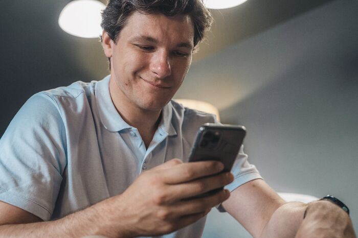 Young man in a light blue shirt reacting with a slight smile to an accidental text message on his smartphone indoors.