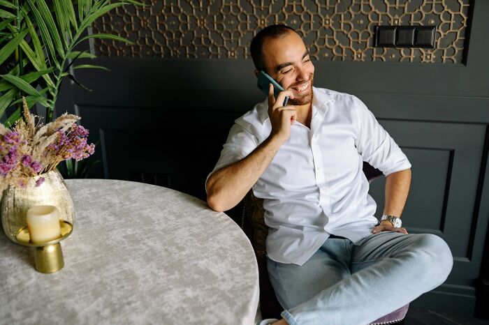 Man smiling while talking on phone, sitting by a round table with flowers, illustrating accidental texts causing panic.