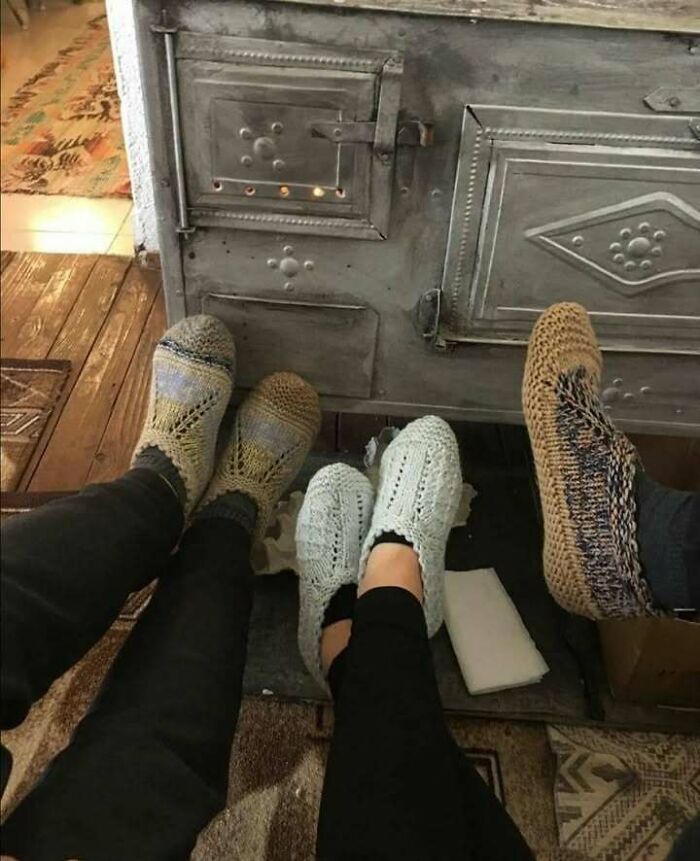 Three people wearing colorful knitted socks warming their feet by an old metal stove, capturing the Eastern European experience.