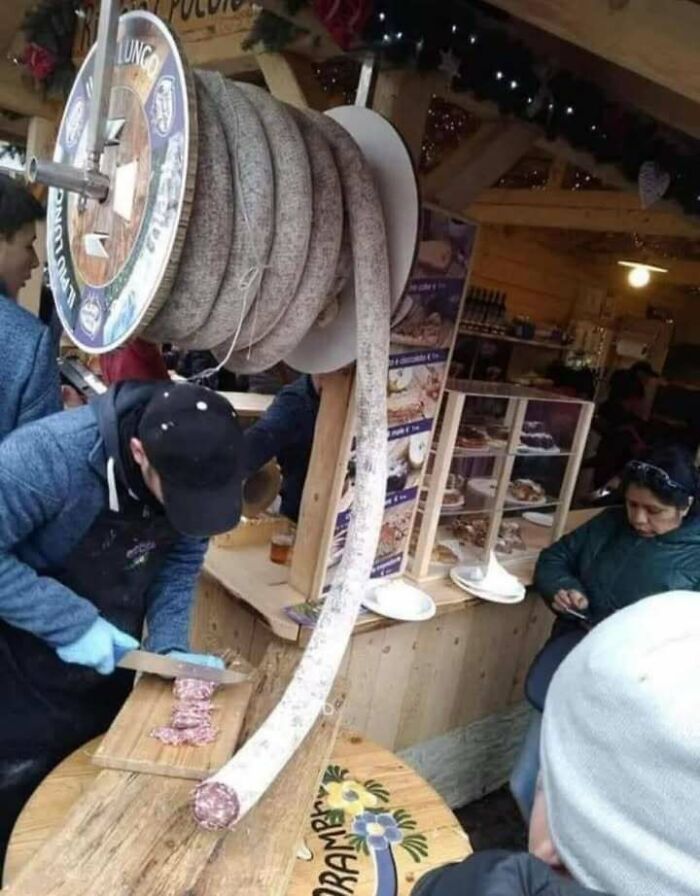 Man slicing a long coil of sausage at a market stall, capturing a funny Eastern European experience moment.