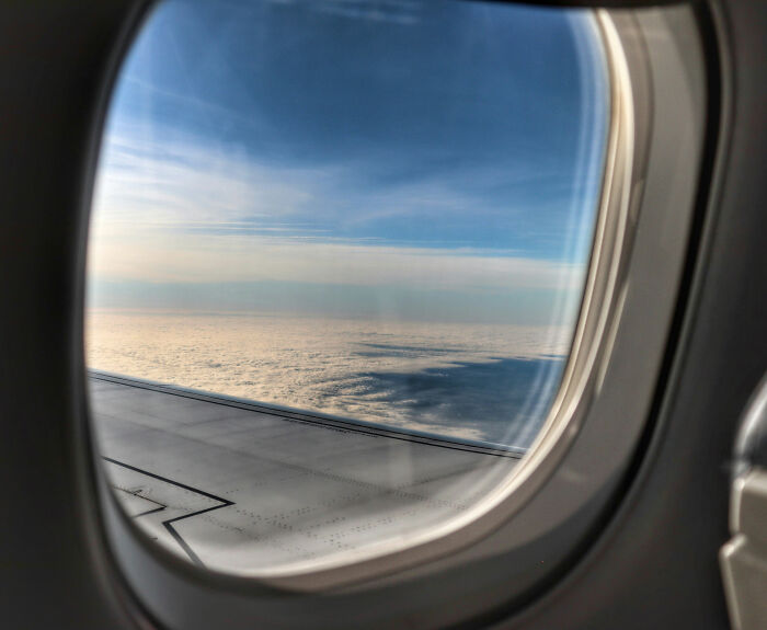 Airplane window view of wing and clouds, passenger highlights sardine can seating and cramped legroom