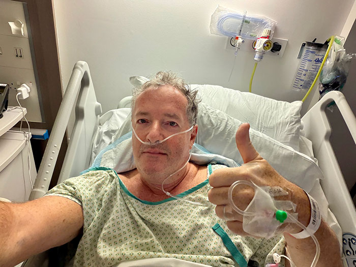 Piers Morgan in hospital bed with IV and oxygen, giving thumbs up after injury involving Donald Trump incident