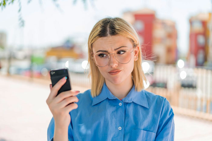 Young woman in glasses looking skeptical at her smartphone while standing outdoors on a sunny day.
