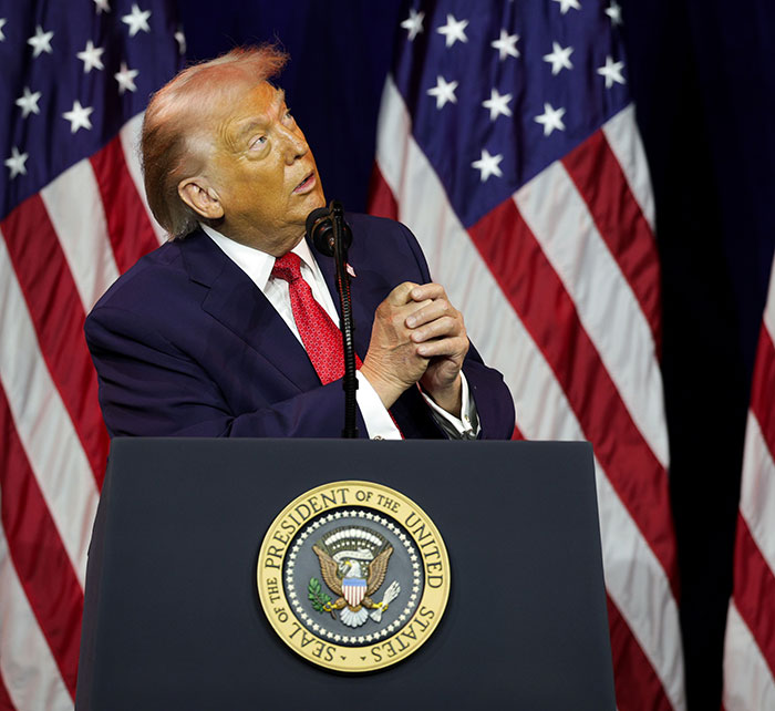 Former President Trump at a podium with multiple American flags, showing hair with a pinkish tint, styled professionally.